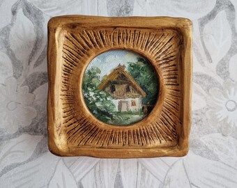 May include: A small, framed painting of a cottage with a thatched roof. The painting is set in a wooden frame with a gold finish.