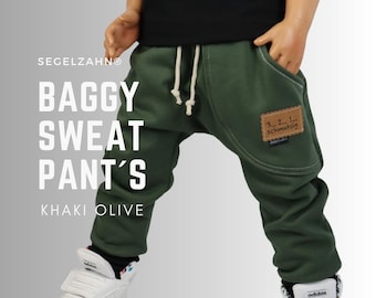 May include: Olive green baggy sweatpants featuring a drawstring waist, deep side pockets, and a rectangular brown patch on the left pocket with the text 3... 2... 1... schmutzig. The pants are paired with a black shirt and white sneakers.