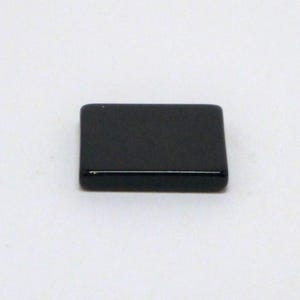 May include: A small, rectangular black pin with a silver-coloured horizontal line near the bottom. The pin is set against a white background. The pin is likely a clothing accessory.
