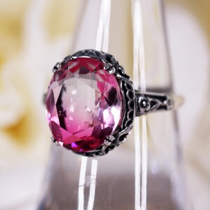 May include: A silver ring with an oval, pink gemstone. The gemstone is the focal point, set in a detailed, dark metal filigree. The ring is displayed on a clear stand against a soft, blurred background.