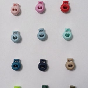 Könnte beinhalten: Sortiment von 15 Kunststoff-Schiebeverschlüssen in verschiedenen Farben, darunter Weiß, Rosa, Rot, Blau, Grün und Schwarz. Die Verschlüsse sind rund und haben einen federbelasteten Mechanismus.