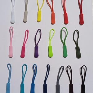 Könnte beinhalten: Sortiment von 20 Reißverschluss-Zierbändern in verschiedenen Farben, darunter Weiß, Grau, Gelb, Orange, Rot, Grün, Lila, Blau, Schwarz und Braun. Die Reißverschluss-Zierbänder sind aus Kunststoff und haben oben eine Schlaufe zum Befestigen an einem Reißverschluss.