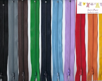 Könnte beinhalten: Eine Sammlung farbiger Reißverschlüsse in verschiedenen Farbtönen, darunter Schwarz, Grau, Braun, Grün, Marineblau, Hellblau, Lila, Rot, Orange und Gelb. Jeder Reißverschluss hat einen Metallschieber und Zähne. Ein Lineal ist sichtbar.