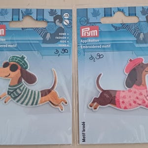 Könnte beinhalten: Zwei Prym Applikationen in transparenter Kunststoffverpackung. Die linke Applikation zeigt einen braunen Dackel mit grüner Baskenmütze, Sonnenbrille und grün gestreiftem Pullover. Die rechte Applikation zeigt einen braunen Dackel mit rosa Baskenmütze und Pullover.