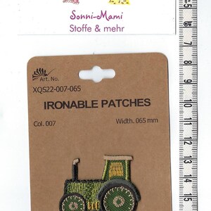 Könnte beinhalten: Bügeleisen-Patch eines grünen Traktors mit gelben Details. Der Patch befindet sich auf einer braunen Karte mit dem Text "Ironable Patches", "Art. No. XQS22-007-065", "Col. 007" und "Width. 0,065 mm".