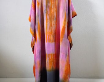 Puede incluir: Una kaftán larga y fluida, de color rosa y naranja con estampado tie-dye y escote en V. La kaftán tiene un dobladillo gris oscuro y está hecha de una tela ligera.