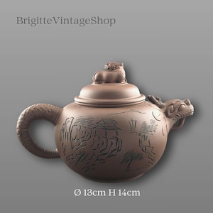 Könnte beinhalten: Braune Keramik-Teekanne mit einem drachenförmigen Ausguss und einem strukturierten Griff. Der Deckel hat eine kleine Tierfigur und gravierte Landschaftsdetails. Durchmesser 13 cm, Höhe 14 cm. Text "BrigitteVintageShop".
