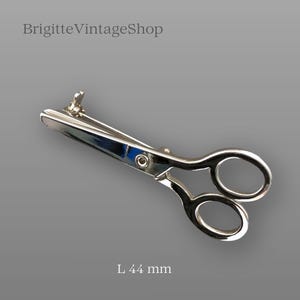 May include: A silver-toned metal brooch shaped like a pair of scissors. The brooch features a polished finish with two circular finger loops and a central pivot point. The text "BrigitteVintageShop" appears at the top, and "L 44 mm" is at the bottom.