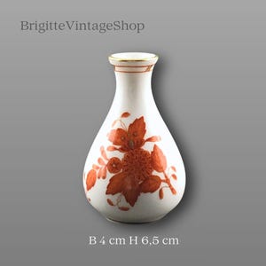 Puede incluir: Jarrón pequeño de cerámica blanca con diseño floral naranja. El jarrón mide 4 cm de ancho y 6,5 cm de alto. El borde está adornado con oro. El texto "BrigitteVintageShop" está en la parte superior izquierda.