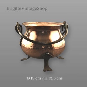 Puede incluir: Un caldero de cobre con textura martillada, con un asa de metal oscuro y tres patas decorativas. El caldero mide aproximadamente 13 cm de diámetro y 12,5 cm de altura. El texto "BrigitteVintageShop" es visible en la parte superior.