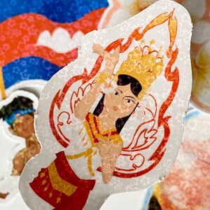 May include: A sticker featuring a colorful illustration of a woman in a traditional Cambodian costume. The woman is standing with her hands clasped in front of her, and she is wearing a red and gold skirt, a white top, and a gold crown. The background of the sticker is white with a red and gold floral pattern.