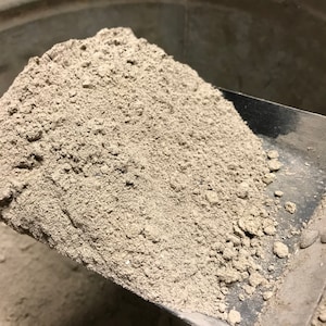 May include: A close-up of a scoop filled with a pile of fine, light gray powder. The powder has a textured, granular appearance. The scoop is made of metal and is partially inside a metal container.