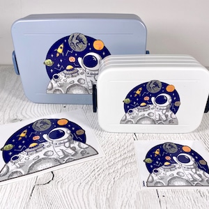 Puede incluir: Dos fiambreras blancas con un diseño espacial azul y blanco que presenta un astronauta, planetas y un cohete. Las fiambreras están sobre una superficie de madera blanca. También hay dos pegatinas con el mismo diseño.