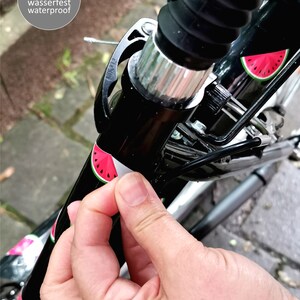 May include: A close-up of a black bicycle handlebar with a watermelon sticker. The sticker is being peeled off to reveal the black handlebar underneath. The sticker is labeled "Sticker wasserfest waterproof". The text "Aufkleber von Jabalou lassen sich r&uuml;ckstandslos entfernen" is visible at the bottom of the image.