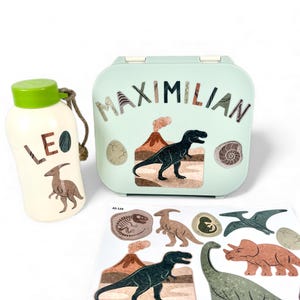 Könnte beinhalten: Ein hellgrünes Lunchbox- und Trinkflaschen-Set mit Dinosaurier-Grafiken und dem Namen "MAXIMILIAN". Die Lunchbox hat einen schwarzen Dinosaurier-Aufkleber und die Trinkflasche einen braunen Dinosaurier-Aufkleber. Ein Bogen mit Dinosaurier-Aufklebern ist ebenfalls enthalten.