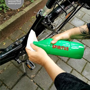 May include: A person cleaning a black bicycle frame with a white cloth and a green bottle labeled "SPIRTIUS". The text "Sticker wasserfest waterproof" is visible in the upper left corner of the image. The text "Die zu beklebenden Fl&auml;chen sollten m&ouml;glichst fettfrei sein" is visible at the bottom of the image.