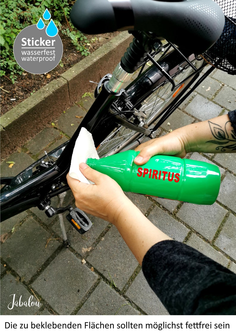 May include: A person cleaning a black bicycle frame with a white cloth and a green bottle labeled "SPIRTIUS". The text "Sticker wasserfest waterproof" is visible in the upper left corner of the image. The text "Die zu beklebenden Fl&auml;chen sollten m&ouml;glichst fettfrei sein" is visible at the bottom of the image.