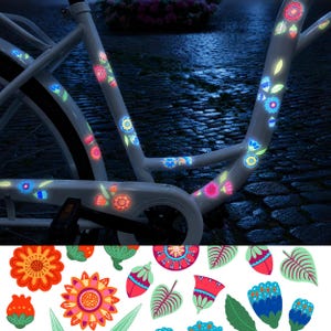 Op de afbeelding: Een wit fietsframe versierd met reflecterende, veelkleurige bloem- en bladstickers die in het donker oplichten. De stickers hebben verschillende vormen, waaronder oranje, roze en blauwe bloemen, samen met groene bladeren en knoppen.