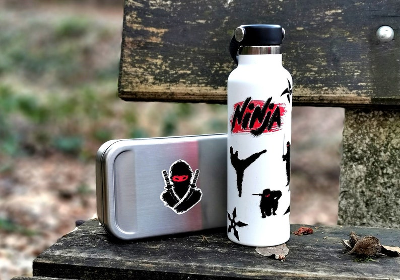 May include: A white water bottle with a black lid and a red and black "Ninja" sticker. The bottle is decorated with black silhouette stickers of ninjas and throwing stars. A silver metal container with a black ninja sticker is next to the bottle.