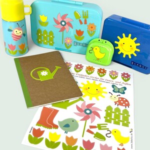 Può includere: Una collezione di lunchbox colorati, un thermos, un taccuino e adesivi con disegni a tema giardino. I lunchbox sono blu e verdi, con fiori, un sole e una girandola. Il thermos è blu e giallo con un'ape e tulipani.