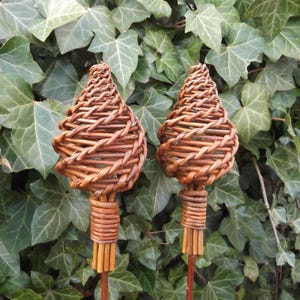 Könnte beinhalten: Zwei dekorative Gartenstecker mit gewebten braunen Weidenholzspitzen in Tropfenform. Die Stecker haben einen zylindrischen Sockel und sind vor einem Hintergrund aus grünen Efeublättern platziert. Ein natürlicher, rustikaler Akzent für Außenbereiche.