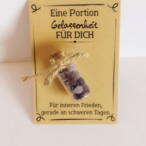 Könnte beinhalten: Eine kleine Glasflasche gefüllt mit lila Edelsteinen, mit Bindfaden gebunden und an einer beigefarbenen Karte befestigt. Die Karte hat deutschen Text: "Eine Portion Gelassenheit FÜR DICH" und "Für inneren Frieden, gerade an schweren Tagen."