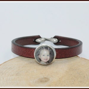 Könnte beinhalten: Ein braunes Lederarmband mit einem silbernen Verschluss und einem runden Foto-Anhänger mit einem Kinderporträt.