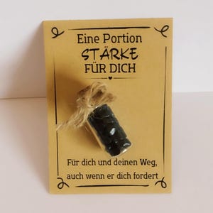 Könnte beinhalten: Eine kleine Glasflasche mit dunklen, zylindrischen Objekten, die mit Bindfaden gebunden ist, ist an einer beigefarbenen Karte befestigt. Die Karte hat dekorative Wirbel und deutschen Text: "Eine Portion Stärke für Dich" und "Für dich und deinen Weg, auch wenn er dich fordert."