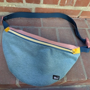 Könnte beinhalten: Eine blaue Cord-Bauchtasche mit einem rosa und gelben Reißverschluss. Die Tasche hat einen marineblauen Riemen und ein kleines schwarzes Etikett mit den Buchstaben "m.i.". Die Tasche liegt auf einer Ziegeloberfläche.