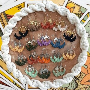 Puede incluir: Una colección de pendientes de macramé hechos a mano exhibidos en una bandeja redonda y tejida. Los pendientes presentan varios diseños, incluyendo soles, lunas y patrones geométricos, con detalles metálicos dorados y plateados. La paleta de colores incluye negro, naranja, verde y morado.