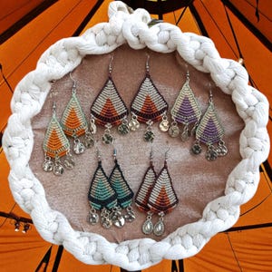 Op de afbeelding: Een verzameling handgemaakte macramé oorbellen in verschillende kleuren, waaronder oranje, teal, paars en bruin. Elke oorbel heeft een driehoekig ontwerp met zilverkleurige bedels. De oorbellen worden gepresenteerd op een beige oppervlak binnen een wit touwframe.