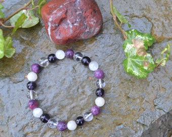 May include: A purple, white, and black beaded bracelet on a gray stone background. The bracelet is made of round beads and has a simple design.