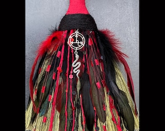 Puede incluir: Una escoba roja, negra y dorada con plumas y cintas. Tiene un amuleto de pentagrama plateado con una serpiente colgando. La escoba está etiquetada como "Lilith Altar Besom" y "Handcrafted By Gypsygold".