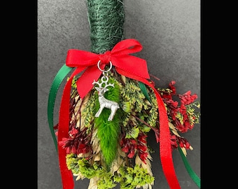 Puede incluir: Un besom de altar de Yule con una cinta roja y verde, un amuleto de reno plateado y flores y hierbas secas.