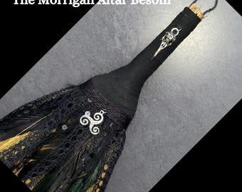 Puede incluir: Besom negro y dorado con encaje negro y plumas. El mango es de tela negra con un amuleto de cuervo plateado y un amuleto de triple espiral plateado. El texto "The Morrigan Altar Besom" y "Handmade By Gypsygold" es visible.
