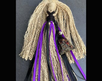 Op de afbeelding: Een handgemaakte beschermingspop met paarse en zwarte linten, een bel en een klein zakje. De pop is gemaakt van natuurlijke vezels en heeft een rustieke, aardse esthetiek. "Protection Poppet" en "Handcrafted By Gypsygold" staan op de afbeelding.