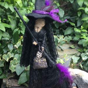 Puede incluir: Una muñeca de bruja negra con un sombrero morado, una cinta morada y una pluma morada. La muñeca sostiene una escoba y una pequeña bolsa de lavanda.