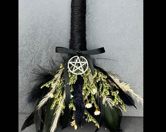 Puede incluir: Una escoba negra con un amuleto de pentagrama, atada con una cinta negra y decorada con flores secas y plumas.