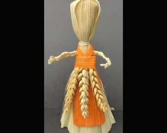 May include: A handmade corn husk doll with an orange skirt and wheat hair. The doll is standing on a gray background. "Corn Dollies" and "Handcrafted By Gypsygold" are written on the image.