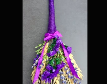 Puede incluir: Una escoba de altar de lavanda hecha a mano. El mango está envuelto en hilo morado, con un lazo y una cinta a juego. La cabeza de la escoba presenta lavanda seca, trigo y vegetación.