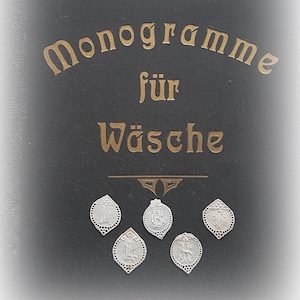 Könnte beinhalten: Ein schwarzer Buchumschlag mit goldfarbenem Schriftzug "Monogramme für Wäsche". Darunter sechs kleine, silberne, dekorative Anhänger mit verschiedenen Monogramm-Designs. Die Anhänger sind rautenförmig mit dekorativem Rand.