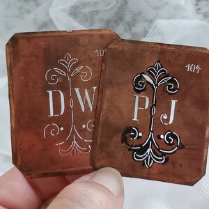 Könnte beinhalten: Zwei Kupferschablonen mit verzierten Designs und den Initialen "DW" und "PJ" in weißer Farbe. Die Schablonen sind mit "10" und "104" nummeriert.