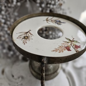 Könnte beinhalten: Ein Vintage-Sockel aus silberfarbenem Metall mit einer runden, weißen Keramikoberfläche. Die Keramikoberfläche weist florale Designs in Braun, Rot und Blau auf. Der Sockel hat ein zentrales Loch und ein dekoratives, gealtertes Aussehen.