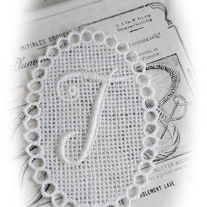 Könnte beinhalten: Weiße Spitzen-Initial "T" mit einem gewellten Rand. Die Spitze liegt auf einem alten Buch mit Text über Stickerei.