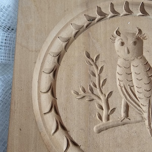 Könnte beinhalten: Eine geschnitzte Holzplakette mit einem Relief einer Eule auf einem Ast in einem kreisförmigen Rahmen. Die Eule hat detaillierte Federn und ist von einem dekorativen Rand umgeben. Das Holz hat eine natürliche, hellbraune Farbe.