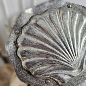 Könnte beinhalten: Ein Vintage-Dekostück aus silberfarbenem Metall in Form einer Muschel. Das Muscheldesign weist strahlenförmige Linien auf und ist in einem achteckigen Rahmen eingefasst. Kleine Nieten befestigen die Muschel am Rahmen. Das Stück hat ein abgenutztes, gealtertes Aussehen.