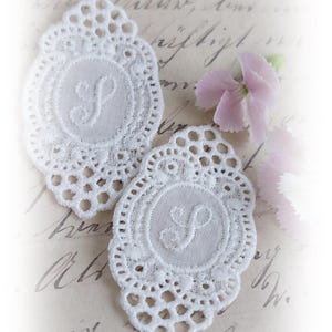 Könnte beinhalten: Zwei weiße Spitzendeckchen mit einem zarten Blumendesign und dem gestickten Buchstaben "S" in der Mitte. Die Deckchen liegen auf einem alten Papierstück mit handschriftlichem Text.
