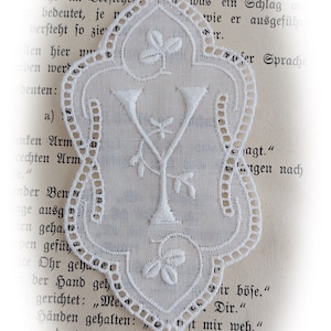 Könnte beinhalten: Weißes, verziertes Spitzenlesezeichen mit floralem und geometrischem Design. Das Lesezeichen befindet sich vor dem Hintergrund alter Buchseiten, wodurch eine Vintage-Ästhetik entsteht. Die Spitze weist filigrane Cutwork-Details auf.