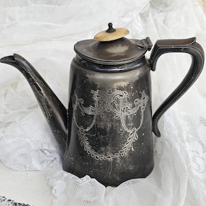 Könnte beinhalten: Eine angelaufene Kaffeekanne aus silberfarbenem Metall mit einer gravierten Schnörkelverzierung auf der Vorderseite, einem geschwungenen Ausguss, einem Klappdeckel mit einem kleinen hellen runden Knopf und einem dunklen Griff. Die Oberfläche zeigt Gebrauchsspuren.