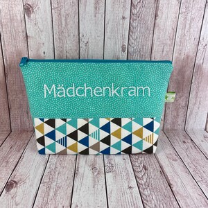 Könnte beinhalten: Eine türkisfarbene Reißverschlusstasche mit einem weißen geometrischen Muster auf der Unterseite und dem aufgestickten Text "Mädchenkram" auf der Vorderseite.